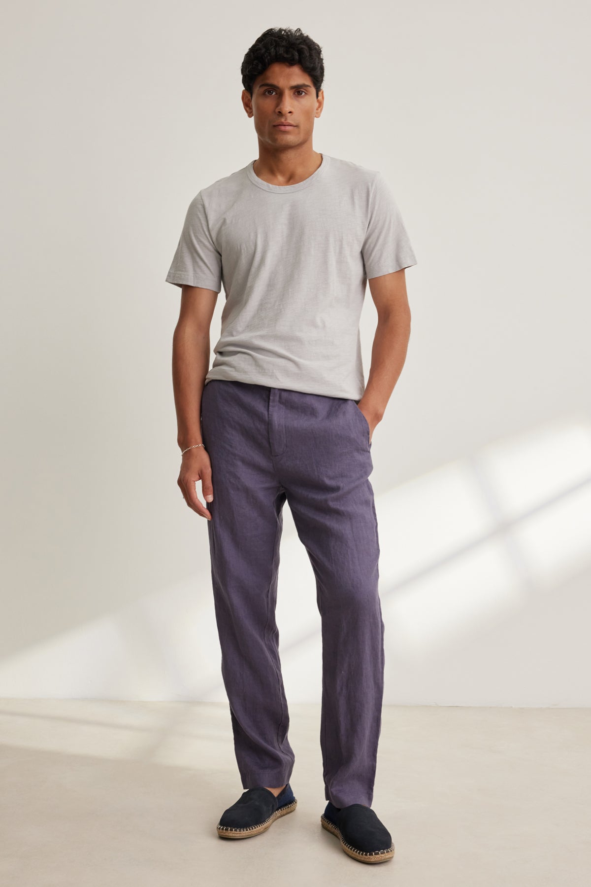 A man stands indoors in a light gray t-shirt and Velvet by Graham & Spencer ALFRED LINEN PANT—purple, breathable, straight-leg with a rear elastic waistband—paired with black slip-on shoes against a plain background.
