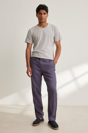 A man stands indoors in a light gray t-shirt and Velvet by Graham & Spencer ALFRED LINEN PANT—purple, breathable, straight-leg with a rear elastic waistband—paired with black slip-on shoes against a plain background.