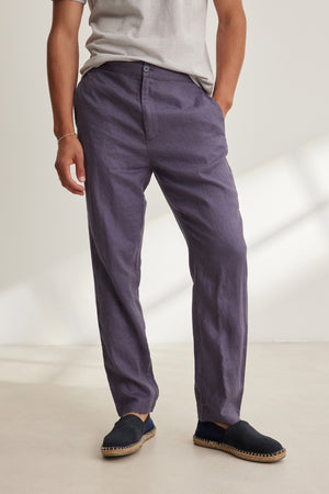 Someone stands indoors with one hand in their pocket, wearing a light grey t-shirt, black espadrilles, and the ALFRED LINEN PANT by Velvet by Graham & Spencer—loose-fitting purple pants featuring an elastic waistband.