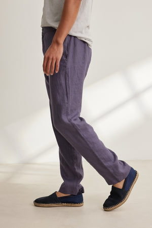 A person stands sideways in a light grey t-shirt and Velvet by Graham & Spencer ALFRED LINEN PANT—breathable, purple, straight-leg pants with an elastic waistband—paired with black slip-on shoes against a neutral background.