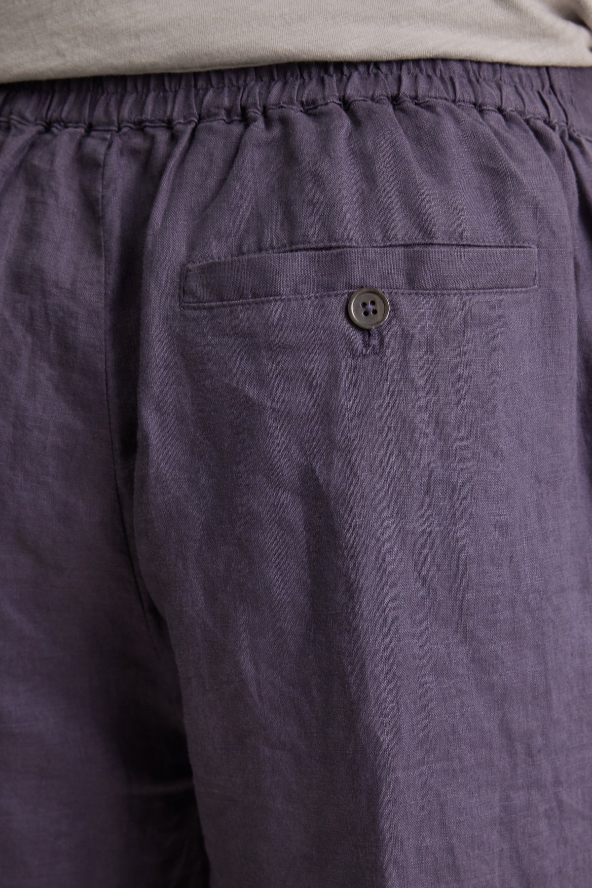 Close-up of the back of a person wearing Velvet by Graham & Spencer's ALFRED LINEN PANT in dark purple, featuring straight legs, breathable linen, a rear elastic waistband, and a buttoned back pocket.
