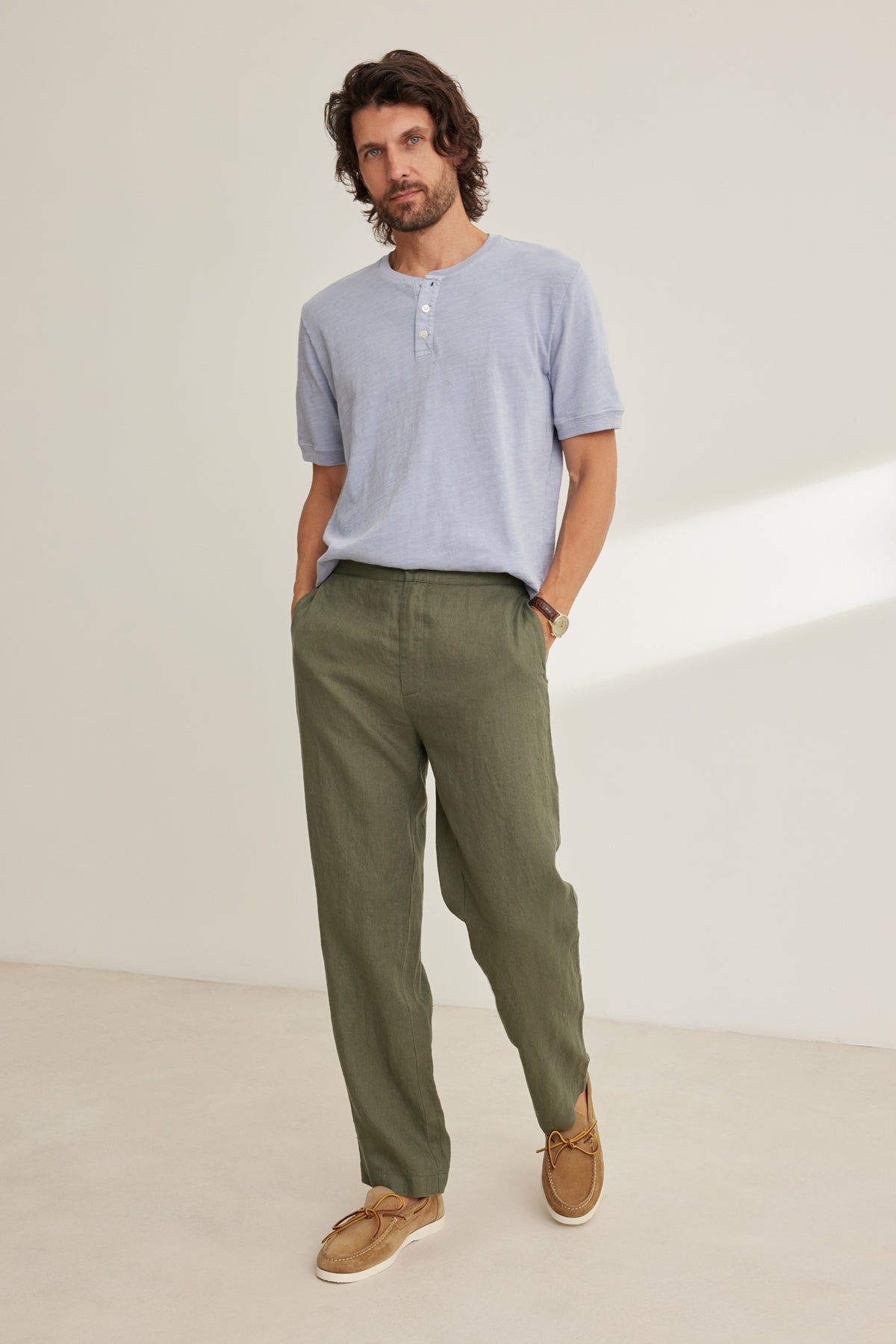 A man stands against a plain background wearing a light blue short-sleeve Henley, the Velvet by Graham & Spencer ALFRED LINEN PANT in olive green with a straight-leg cut, and tan boat shoes, his hands casually in his pockets.-39152330768577
