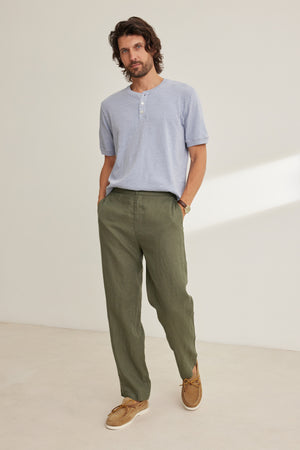 A man stands against a plain background wearing a light blue short-sleeve Henley, the Velvet by Graham & Spencer ALFRED LINEN PANT in olive green with a straight-leg cut, and tan boat shoes, his hands casually in his pockets.