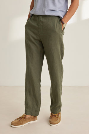 A person stands with hands in pockets against a plain background, wearing Velvet by Graham & Spencer's ALFRED LINEN PANT in olive green with a straight-leg fit and elastic waistband, paired with a light blue shirt and tan boat shoes.