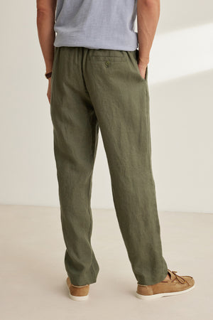 A person stands indoors with hands in pockets, wearing Velvet by Graham & Spencer's ALFRED LINEN PANT in olive green, featuring an elastic waistband and straight-leg cut, paired with a light gray shirt and tan shoes. Back view shown.