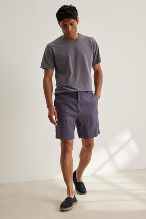 A man stands indoors against a plain white wall, wearing a grey t-shirt and Velvet by Graham & Spencer’s EAMON LINEN SHORT—purple, lightweight with an elastic waistband—ideal for warm weather.