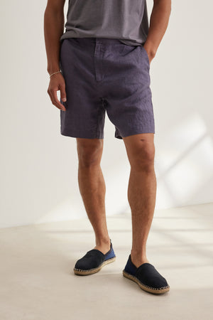 A person indoors wears a grey tee, Velvet by Graham & Spencer's EAMON LINEN SHORT with elastic waistband in dark grey, and black espadrilles—perfect for warm days.