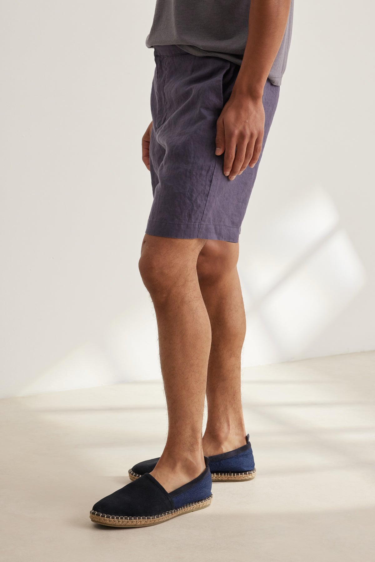 Wearing a grey linen tee, Velvet by Graham & Spencer's EAMON LINEN SHORT in purple, and navy espadrilles, a person stands indoors on a light floor—perfectly dressed for warm weather.