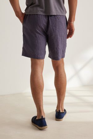 A person stands indoors facing away, wearing a lightweight grey linen tee, Velvet by Graham & Spencer’s EAMON LINEN SHORT in dark grey with an elastic waistband, and navy espadrilles—ideal for warm weather comfort.