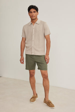 A man stands indoors against a plain light background wearing Velvet by Graham & Spencer's EAMON LINEN SHORT, paired with a beige linen short-sleeve shirt and tan boat shoes—a versatile, stylish look for warm weather.