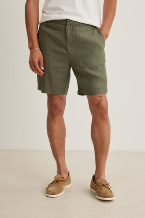 A person wearing the EAMON LINEN SHORT by Velvet by Graham & Spencer, paired with a white tee and brown boat shoes, stands hands in pockets against a plain background, highlighting a versatile warm-weather look.