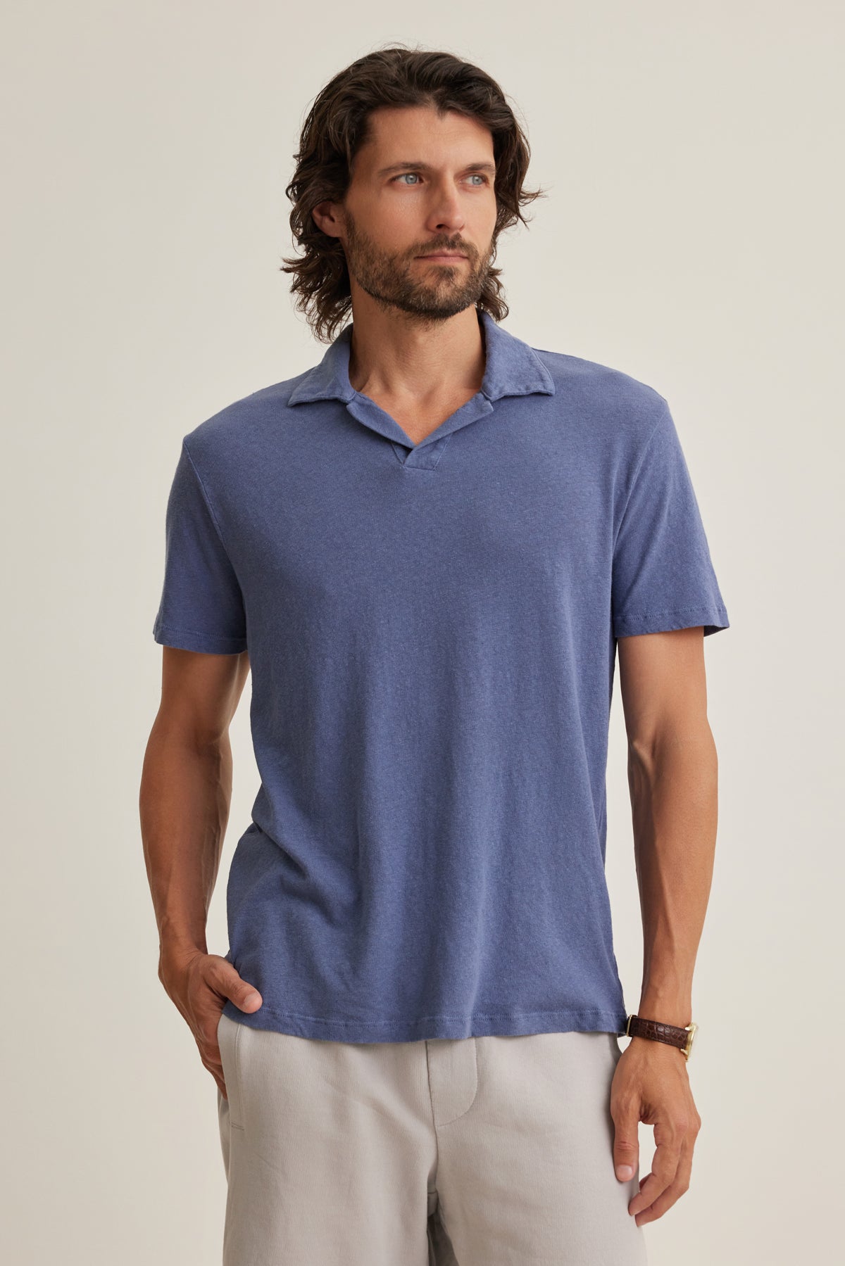 A man with medium-length brown hair and a beard wears the BECK LINEN COTTON POLO by Velvet by Graham & Spencer—a blue, breathable short-sleeve collared shirt—paired with light pants, standing against a plain light background.-39152336502977