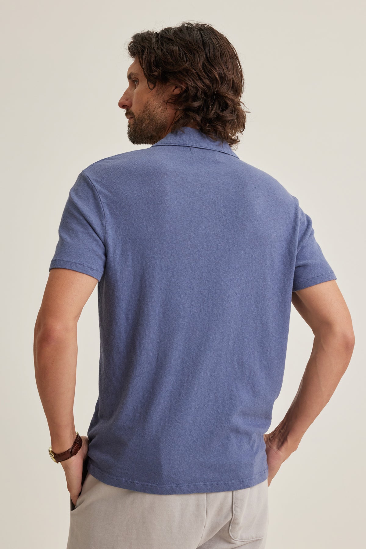 A man stands with his back to the camera, wearing Velvet by Graham & Spencer's BECK LINEN COTTON POLO in blue and light linen-blend pants—an ideal, stylish look for warm-weather days against a plain background.-39152336437441