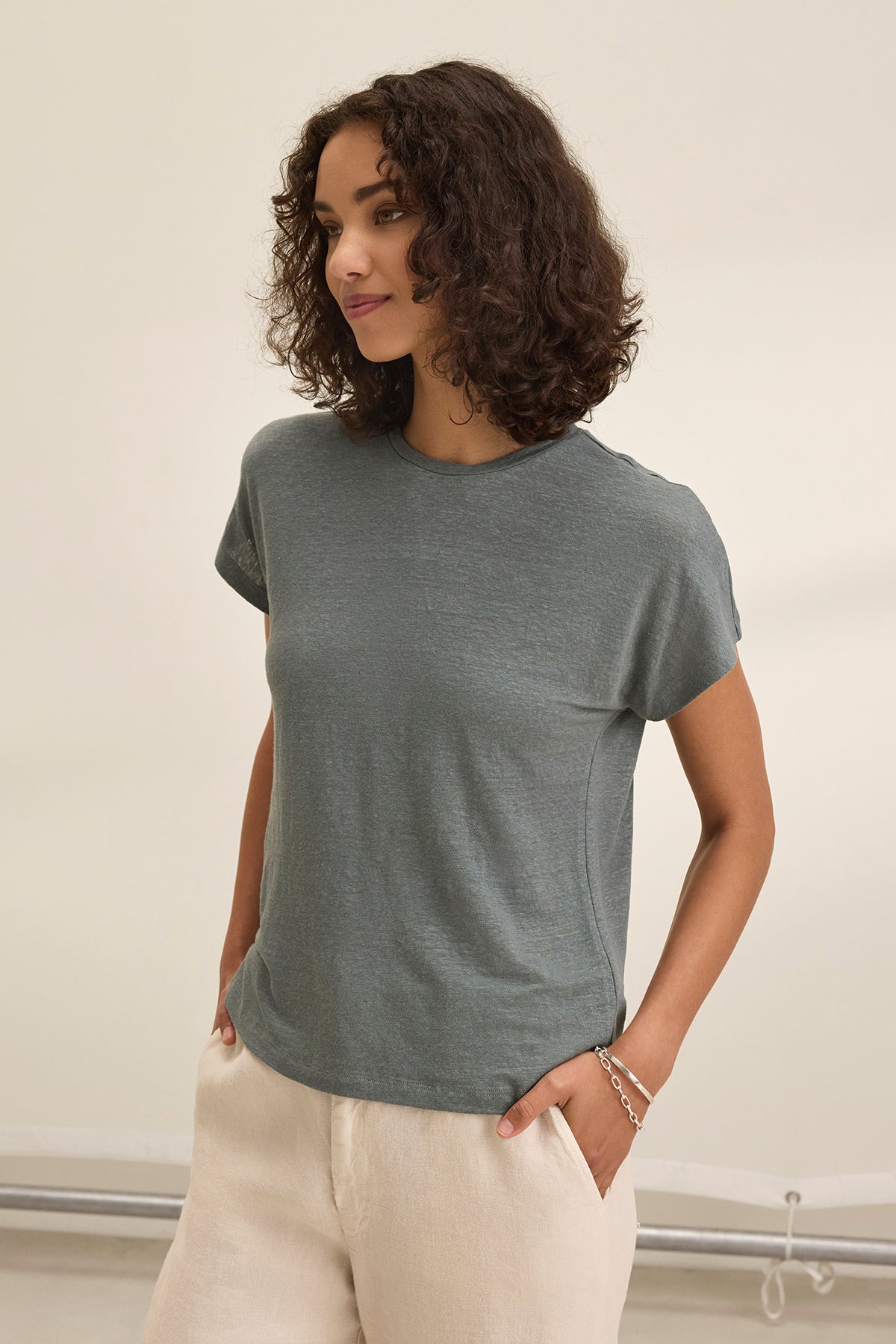 A person with curly hair is wearing a gray JASMINE LINEN KNIT TEE by Velvet by Graham & Spencer and beige, breathable linen knit pants, standing against a neutral background.-38662922895553
