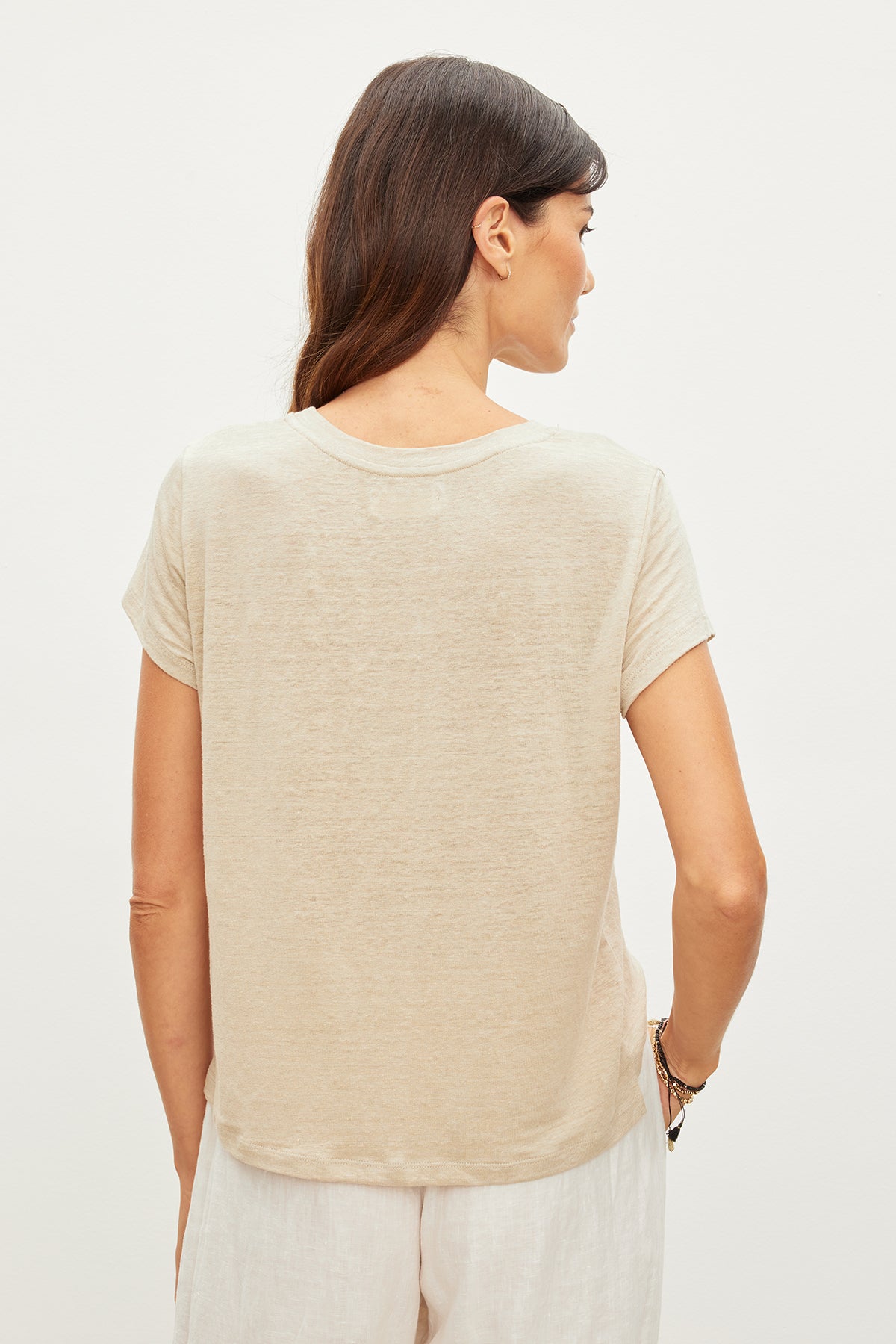 A person with long hair is wearing a CASEY LINEN KNIT TEE by Velvet by Graham & Spencer in beige, paired with white pants, viewed from the back against a plain background.-35955505955009