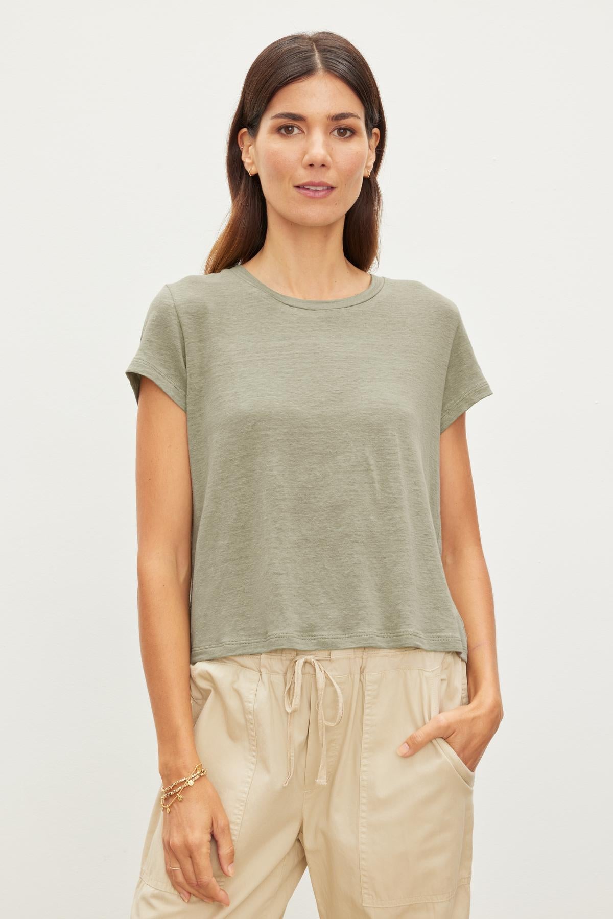 A woman wearing a green Velvet by Graham & Spencer CASEY LINEN KNIT CREW NECK TEE and beige drawstring pants stands against a plain background, looking directly at the camera.