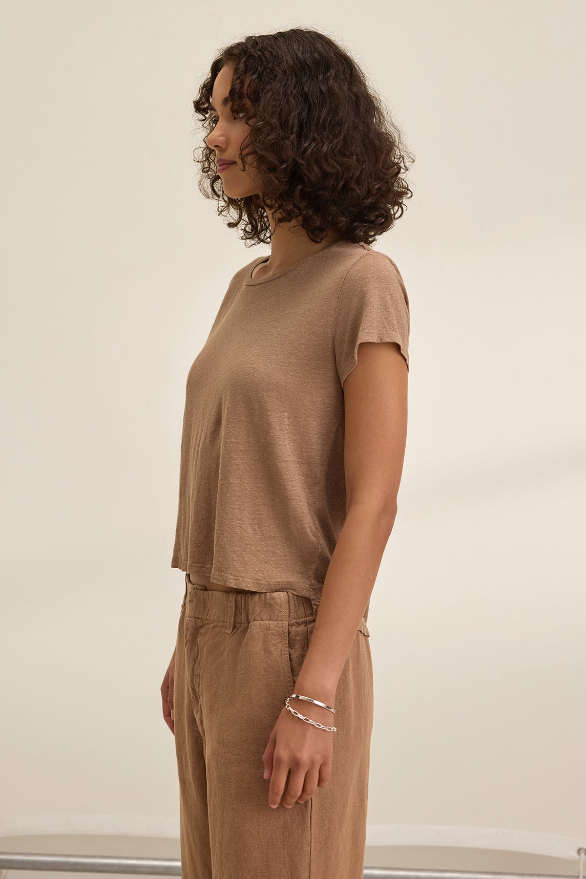A person with curly hair in the CASEY LINEN KNIT TEE by Velvet by Graham & Spencer and brown pants stands in profile against a plain background, highlighting how well the top complements their simple outfit.-38662914670785