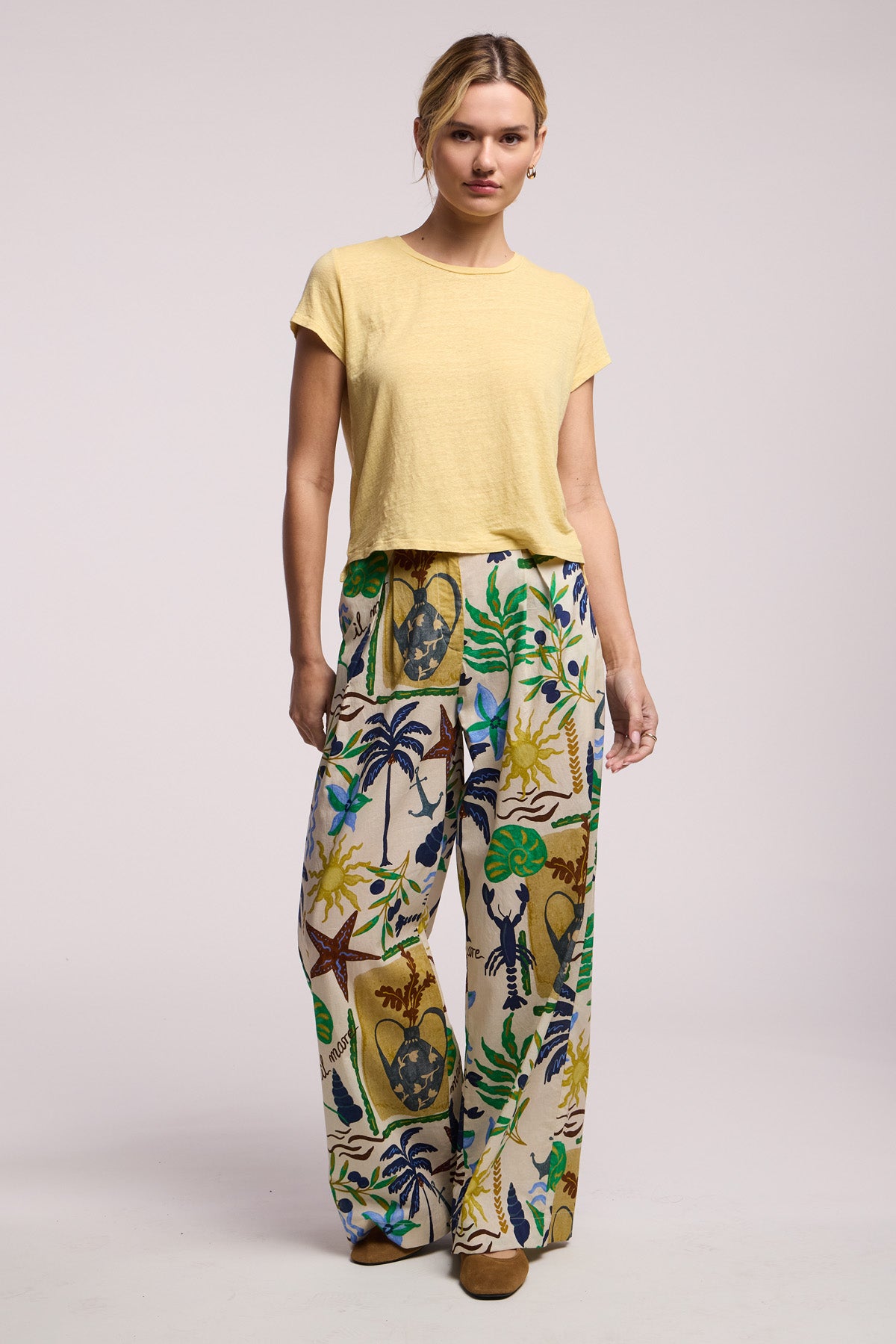 A woman wears the CASEY TEE by Velvet by Graham & Spencer, a short-sleeved yellow linen knit top, paired with wide-leg, high-waisted pants featuring a colorful tropical print, standing against a plain light background.-41526949150913