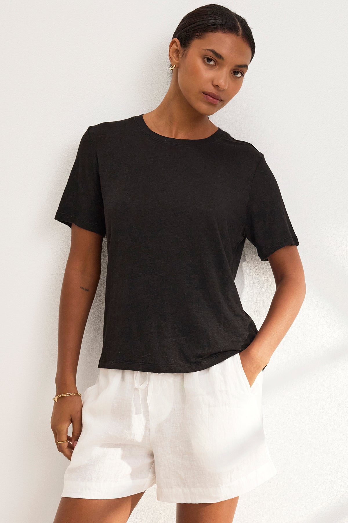 A person stands against a white wall wearing the HERITAGE LINEN KNIT TEE by Velvet by Graham & Spencer in black with white shorts, one hand in their pocket.-39273975185601