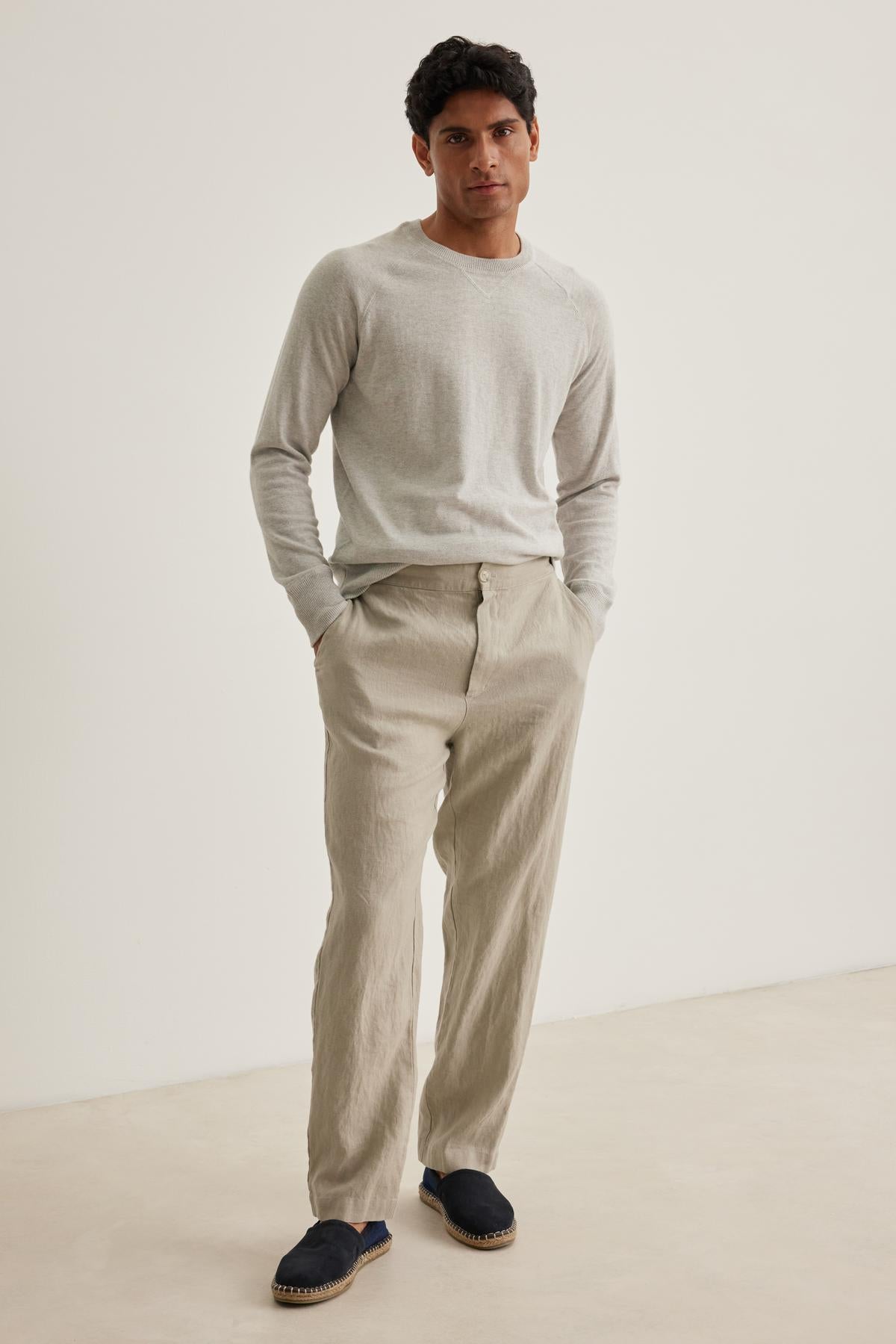 A man in a light gray sweater and Velvet by Graham & Spencer's LENNON LINEN PANT stands against a plain background, hands in pockets, wearing dark slip-on shoes.