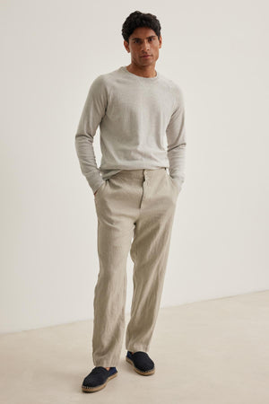 A man in a light gray sweater and Velvet by Graham & Spencer's LENNON LINEN PANT stands against a plain background, hands in pockets, wearing dark slip-on shoes.