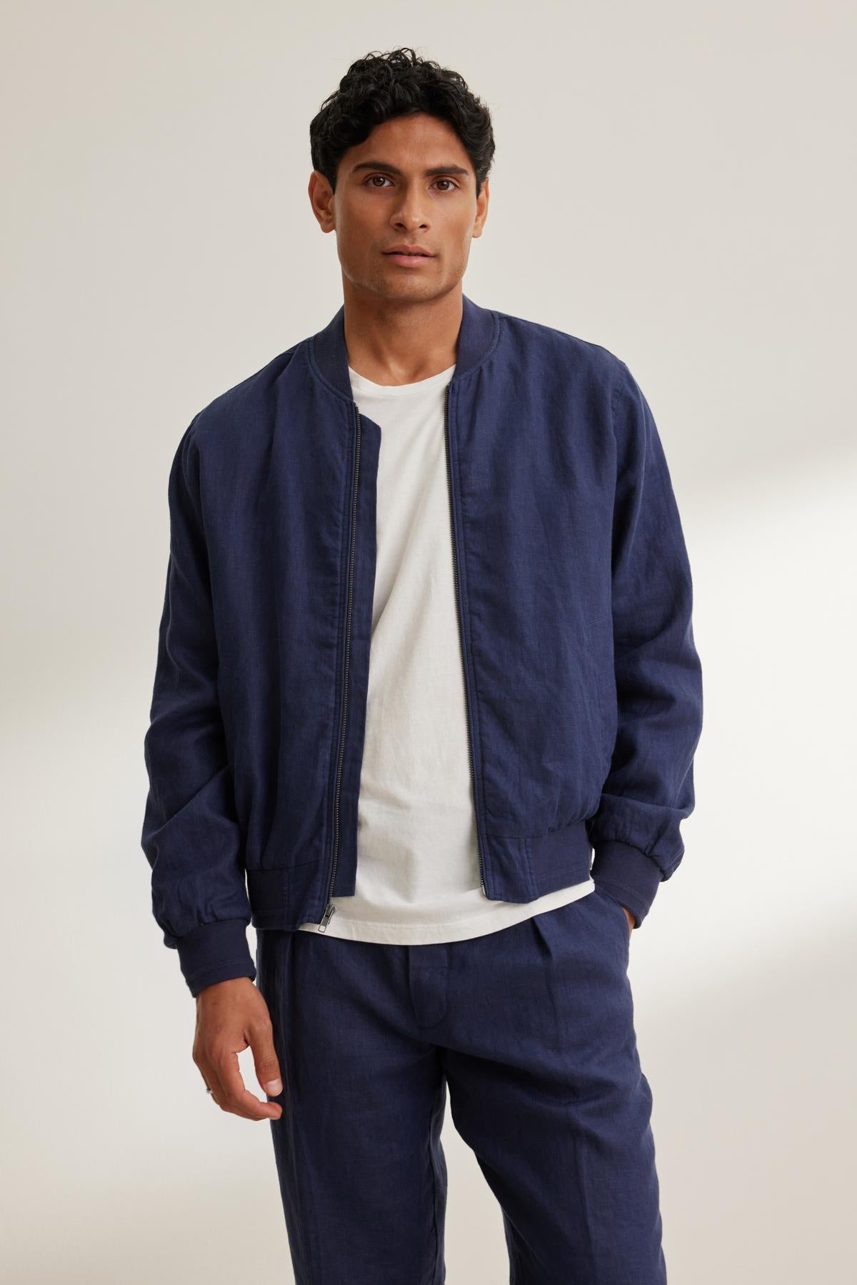 A person wearing a Velvet by Graham & Spencer MURPHY LINEN BOMBER JACKET in navy blue over a white shirt with matching navy pants stands against a plain background.
