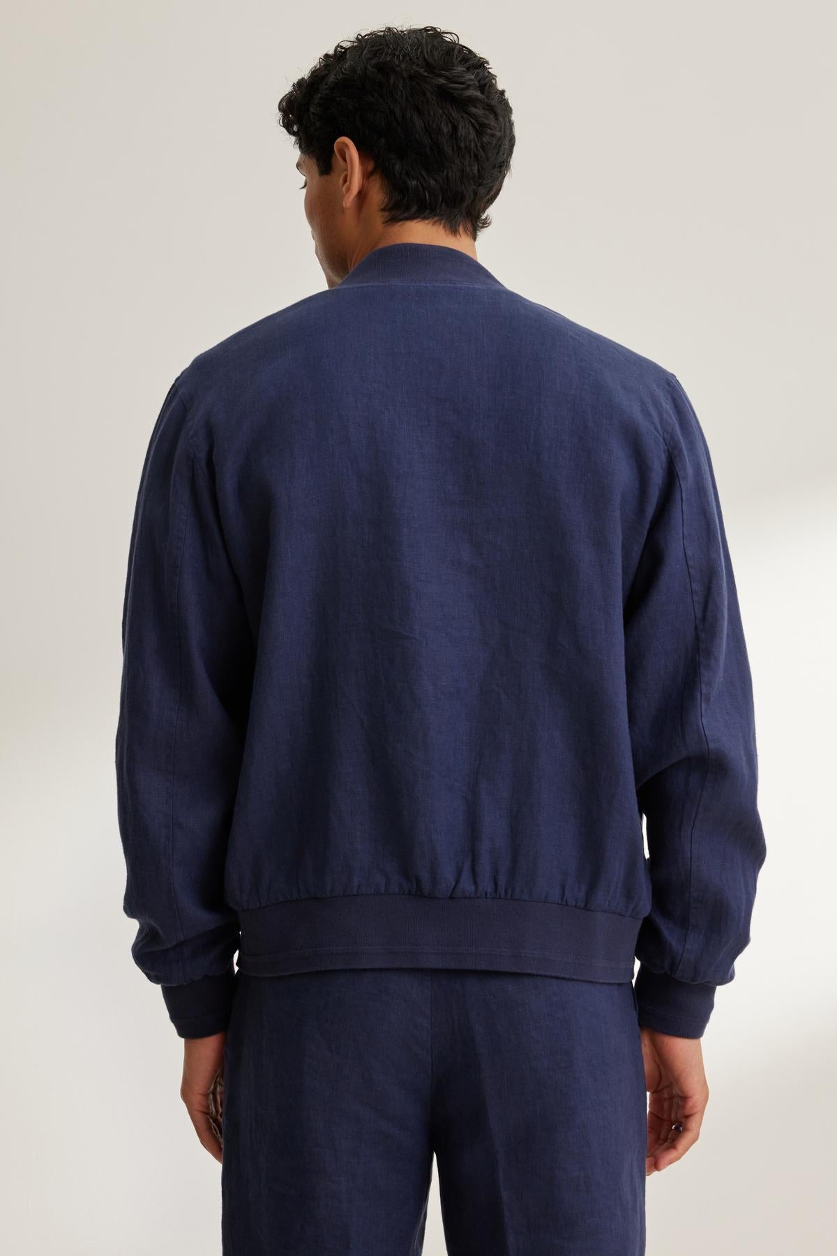 A person with short dark hair wearing a Velvet by Graham & Spencer Murphy Linen Bomber Jacket and dark pants is standing, facing away.