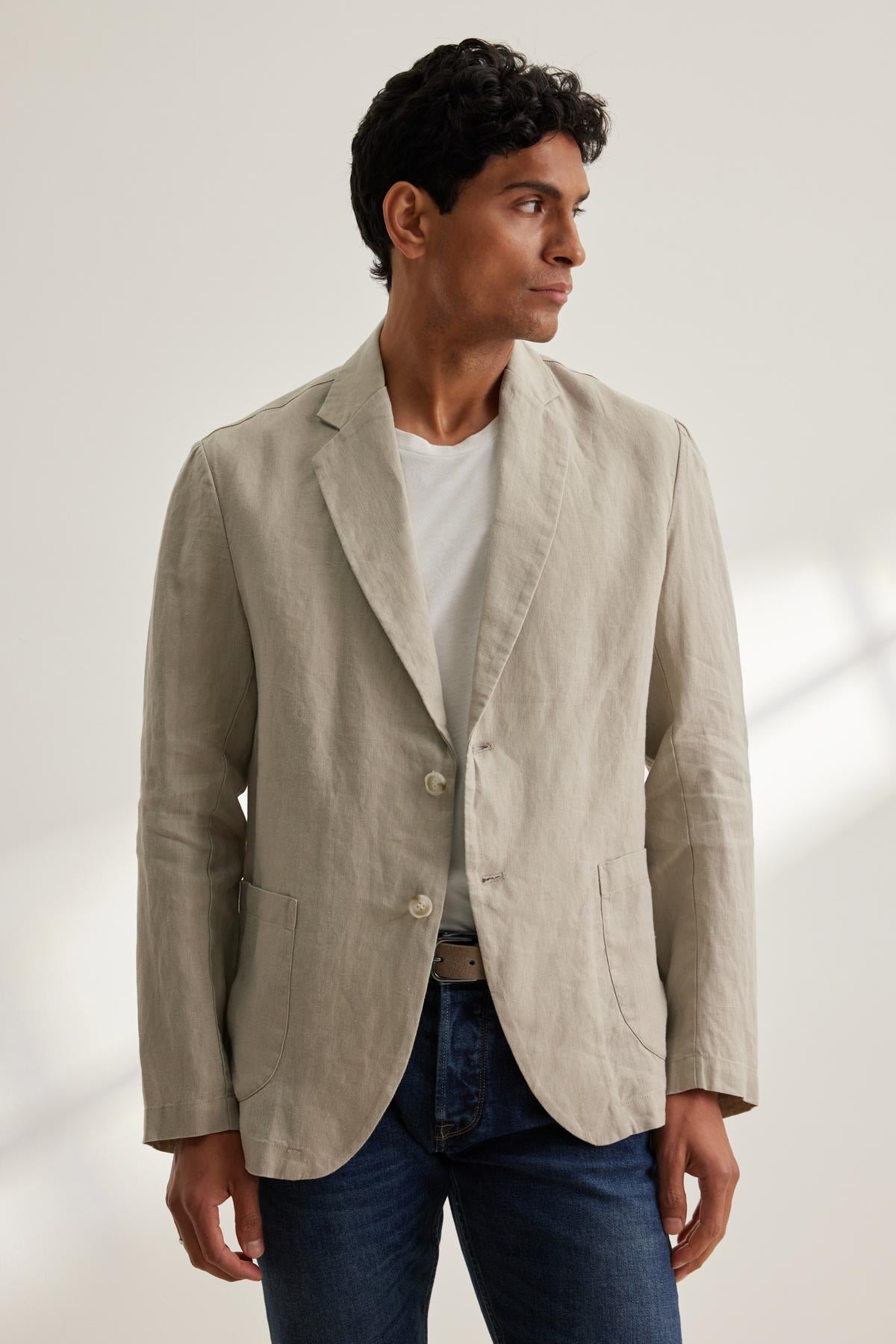 A man wearing the WARREN LINEN BLAZER by Velvet by Graham & Spencer over a white shirt and blue jeans looks to the side.