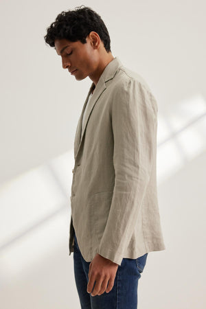 A man in a light beige WARREN LINEN BLAZER by Velvet by Graham & Spencer and jeans stands against a neutral background, gazing thoughtfully downward.