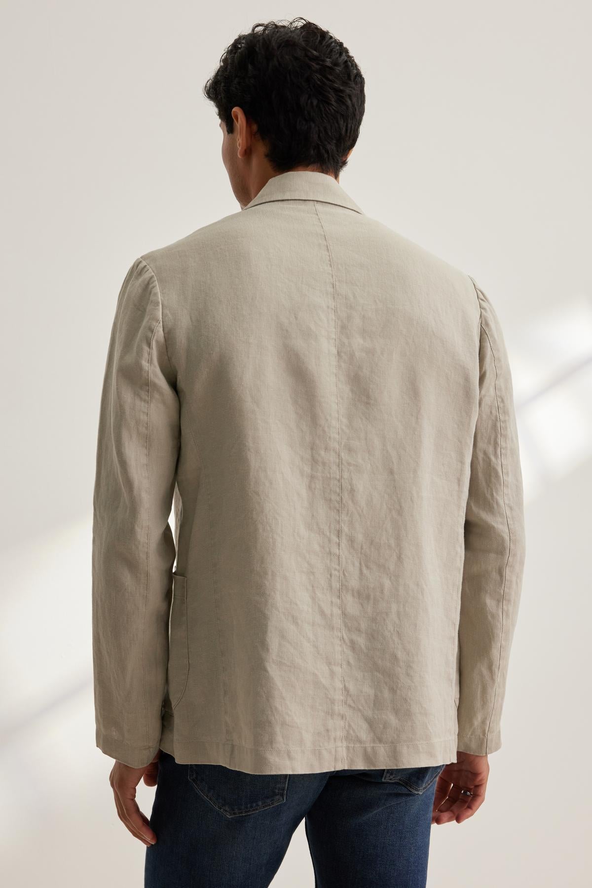 Someone in a WARREN LINEN BLAZER by Velvet by Graham & Spencer and jeans stands facing away in soft lighting.