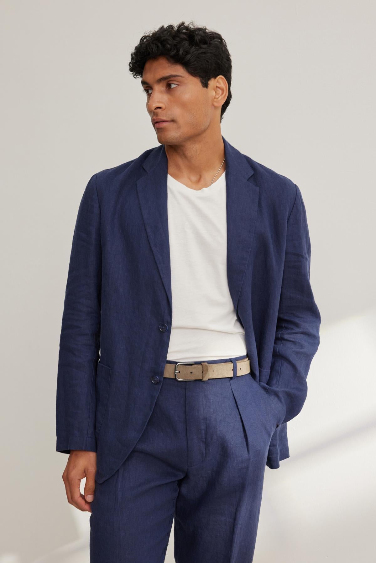 A man in a Velvet by Graham & Spencer WARREN LINEN BLAZER and a white t-shirt stands against a plain background, one hand in his pocket.-38643372392641