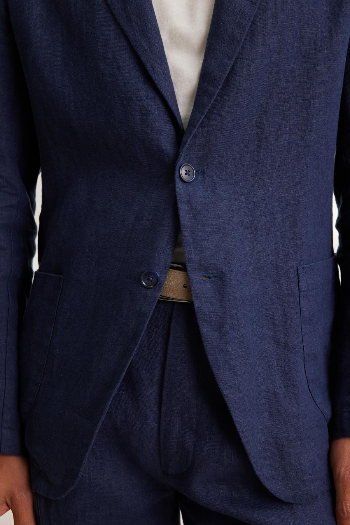 Someone is wearing a WARREN LINEN BLAZER by Velvet by Graham & Spencer in navy blue, paired with a white shirt. The blazer is buttoned, revealing a beige belt partially visible underneath.