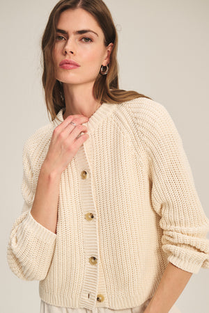 A woman wears the LISABETH CARDIGAN by Velvet by Graham & Spencer—a cream, long-sleeve Pima cotton button-up—against a neutral background, offering effortless style as a perfect transitional layer.