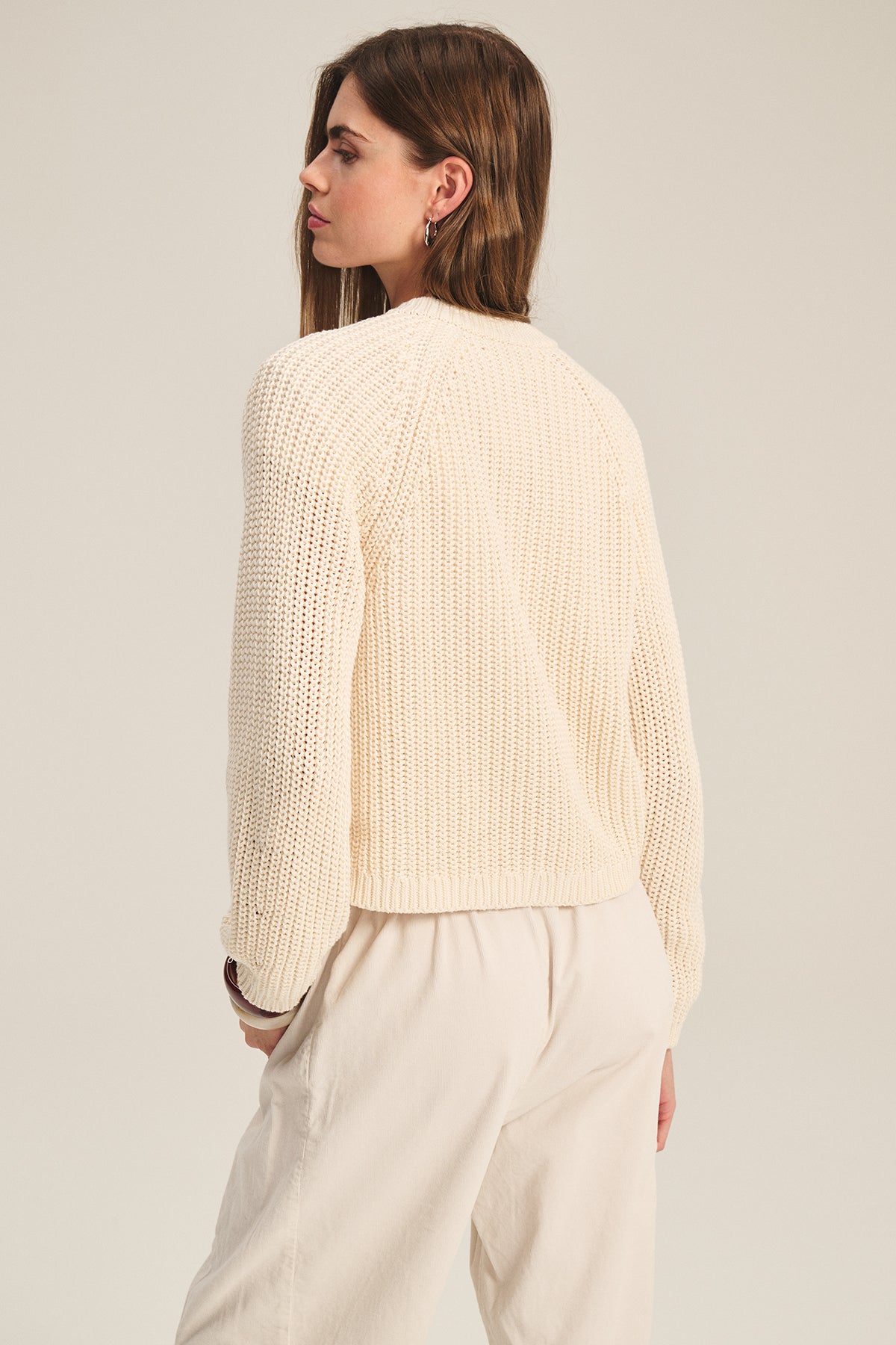 A woman faces away in the Velvet by Graham & Spencer LISABETH CARDIGAN, a chunky cropped cardigan ideal as a transitional layer, paired with light pants against a plain background.