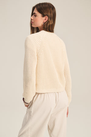 A woman faces away in the Velvet by Graham & Spencer LISABETH CARDIGAN, a chunky cropped cardigan ideal as a transitional layer, paired with light pants against a plain background.
