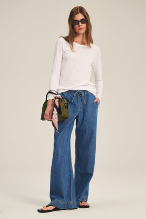 A woman wears the LIZZIE TEE by Velvet by Graham & Spencer, paired with wide-leg blue jeans, sandals, sunglasses, and holds a green handbag with a scarf.