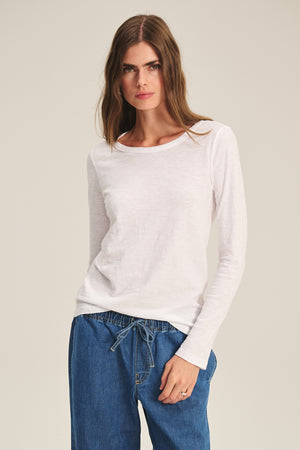 Woman wearing the LIZZIE TEE by Velvet by Graham & Spencer—a plain white long sleeve crewneck—paired with blue drawstring pants, standing against a neutral background.