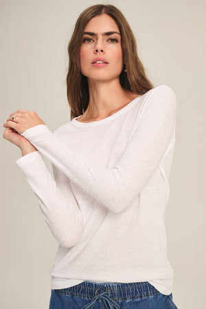 A woman models the LIZZIE TEE by Velvet by Graham & Spencer, featuring long sleeves and a crew neckline, paired with blue pants as she stands against a plain light background, looking at the camera with her arms slightly crossed.