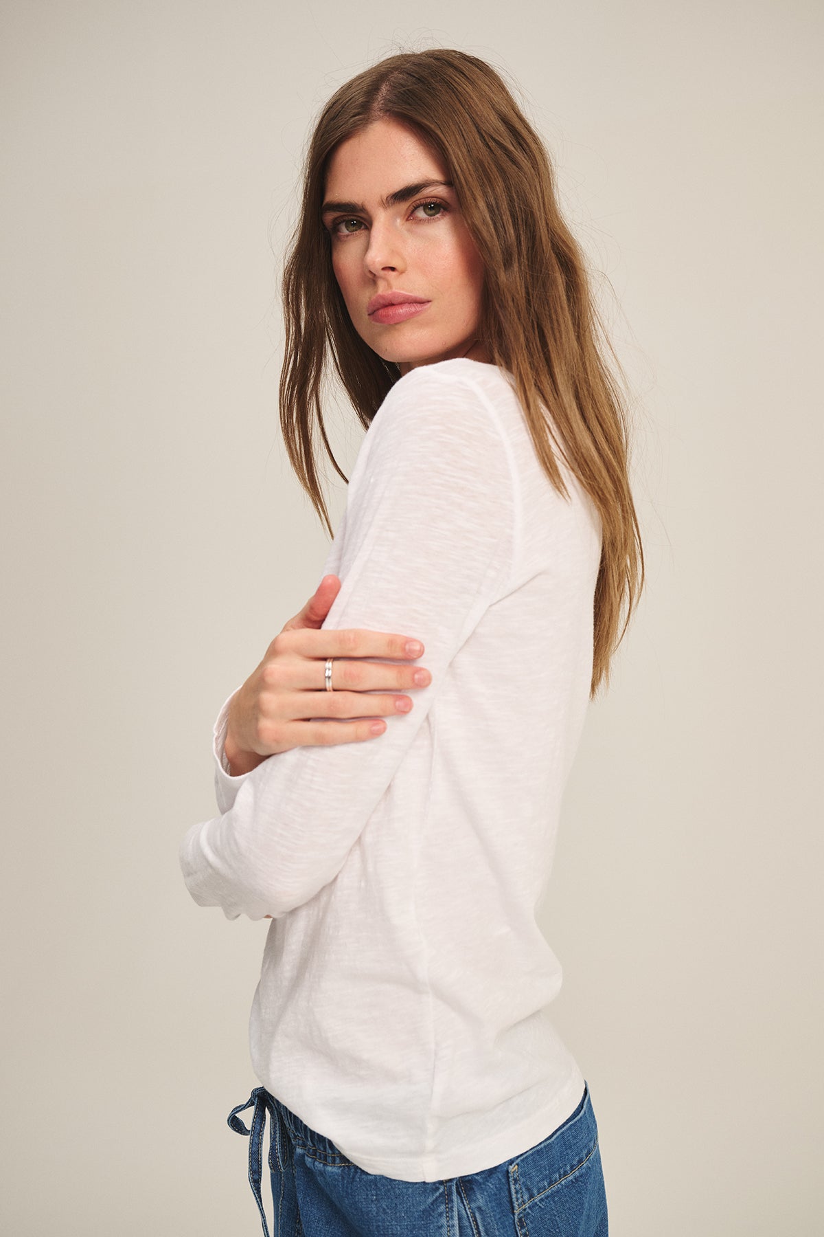 A person with long brown hair wears the LIZZIE TEE by Velvet by Graham & Spencer—a white textured cotton slub long sleeve top—paired with blue jeans, standing with one arm crossed, looking over their shoulder at the camera.