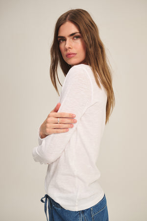 A person with long brown hair wears the LIZZIE TEE by Velvet by Graham & Spencer—a white textured cotton slub long sleeve top—paired with blue jeans, standing with one arm crossed, looking over their shoulder at the camera.