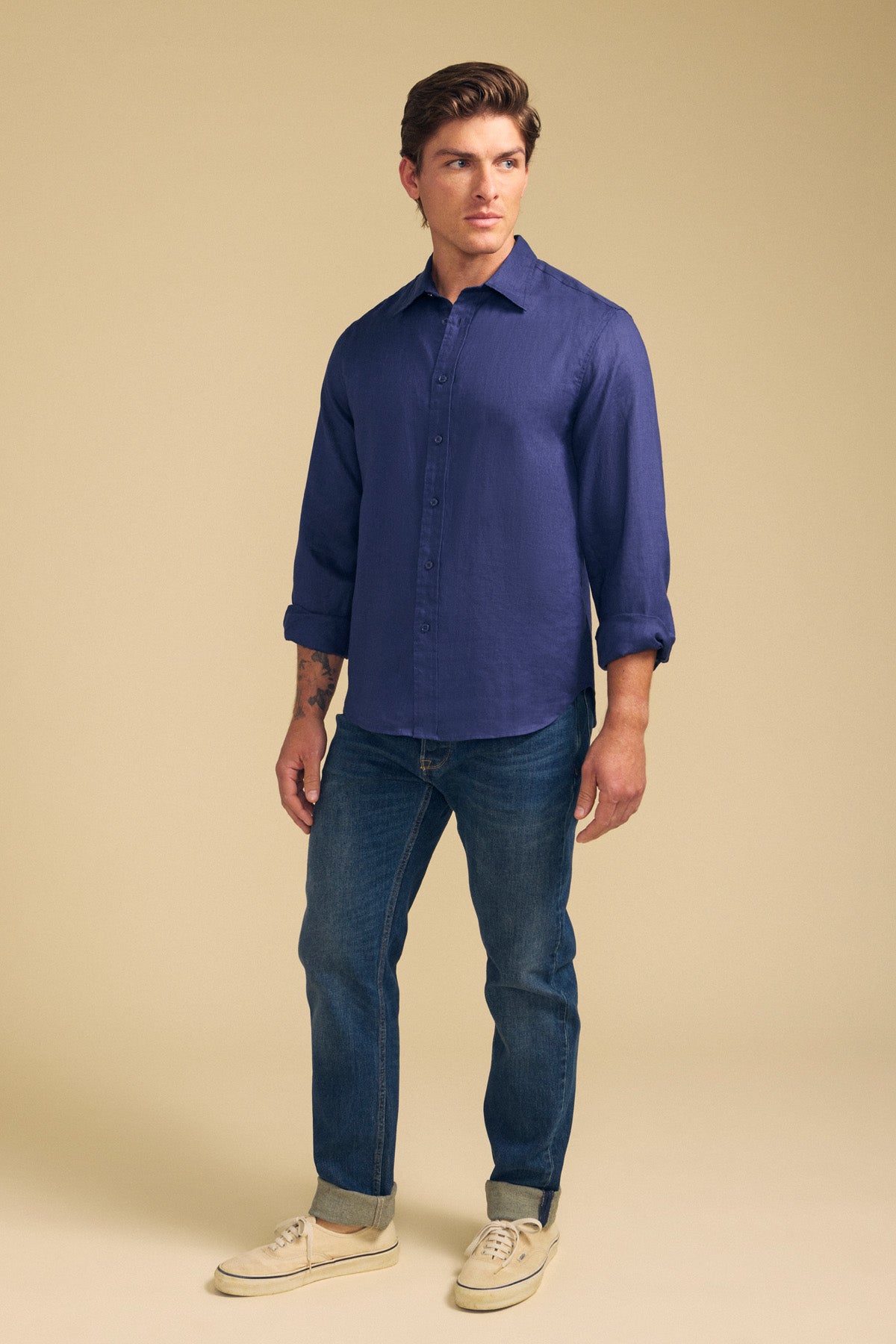 A man wears the CARUSO LINEN SHIRT by Velvet by Graham & Spencer, styled with blue jeans and light sneakers against a plain beige background—a perfect choice for effortless layering.-41701813289153