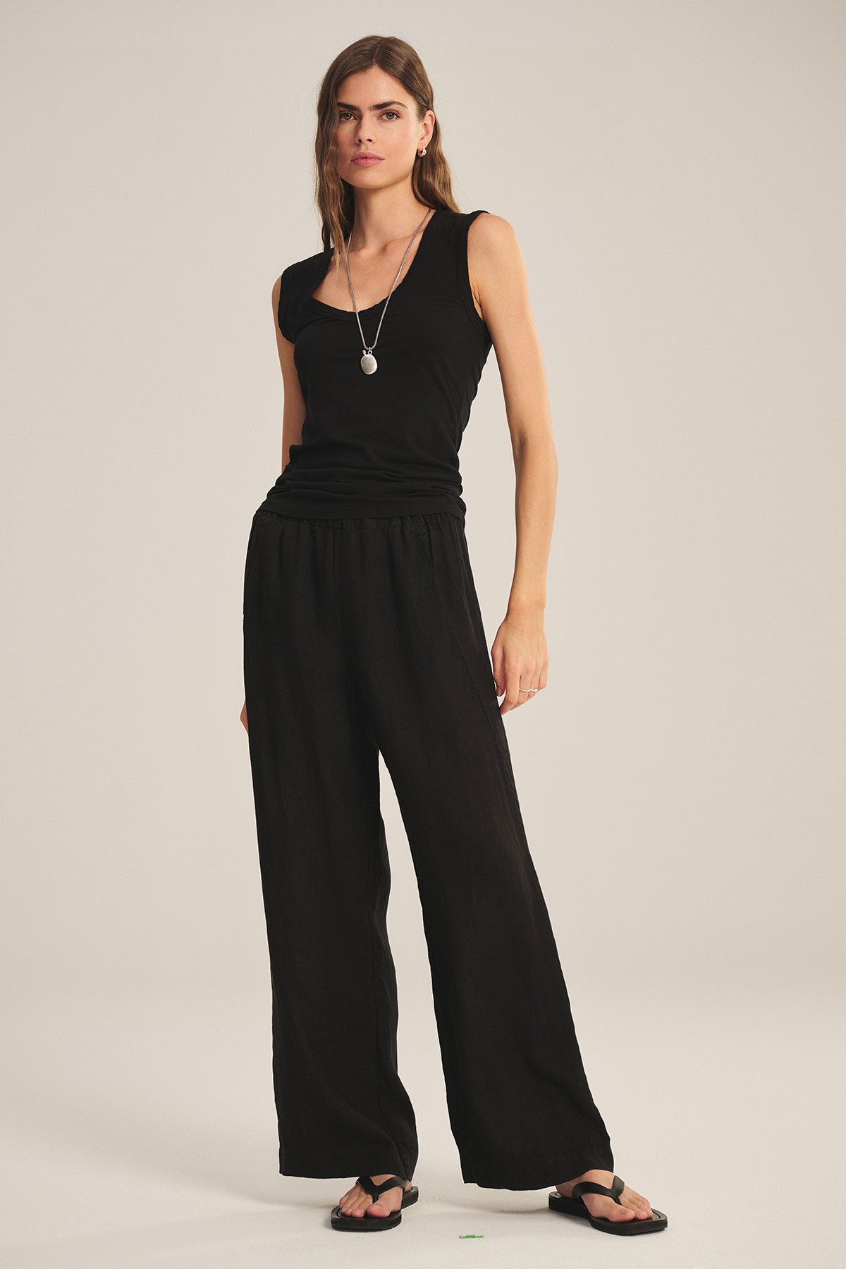 A woman stands against a plain light background wearing Velvet by Graham & Spencer's LOLA LINEN PANT, a sleeveless black top, black sandals, and a necklace.