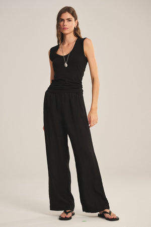 A woman stands against a plain light background wearing Velvet by Graham & Spencer's LOLA LINEN PANT, a sleeveless black top, black sandals, and a necklace.