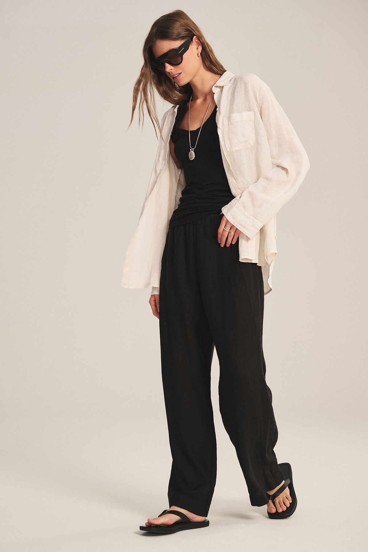 A person stands against a plain background wearing Velvet by Graham & Spencer's LOLA LINEN PANT in black, paired with a black tank top, an open beige shirt, black sandals, sunglasses, and a necklace.