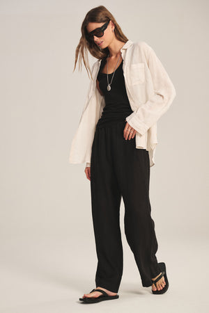 A person stands against a plain background wearing Velvet by Graham & Spencer's LOLA LINEN PANT in black, paired with a black tank top, an open beige shirt, black sandals, sunglasses, and a necklace.
