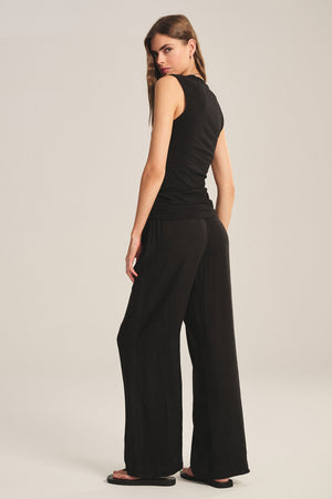 A woman stands facing away, looking over her shoulder. She wears Velvet by Graham & Spencer's LOLA LINEN PANT with a sleeveless black top and black sandals against a plain background.