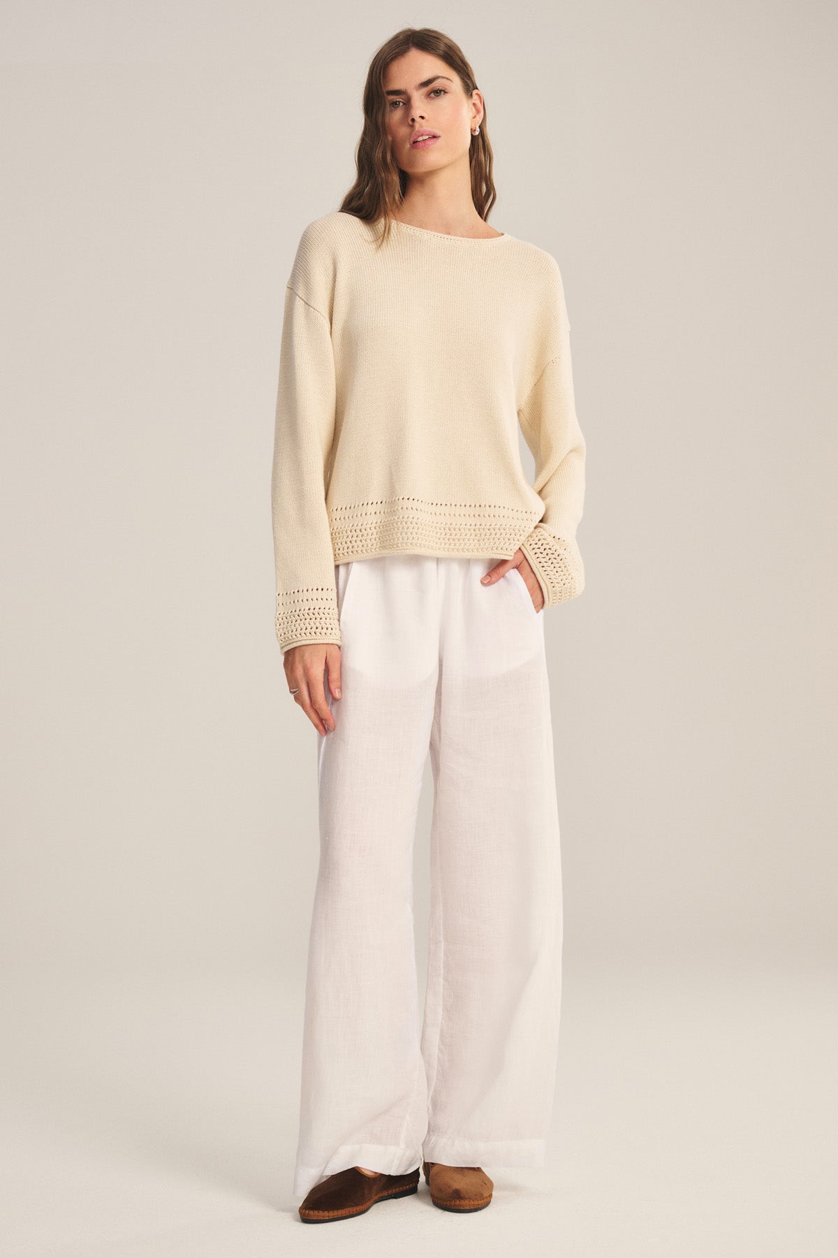 A person stands against a plain background wearing a loose beige sweater with crocheted cuffs and hem, and the LOLA LINEN PANT in white by Velvet by Graham & Spencer, paired with brown shoes.-40828612182209
