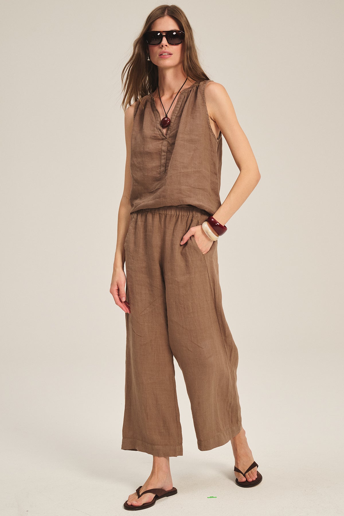 A woman wearing sunglasses and the TACY LINEN TANK TOP by Velvet by Graham & Spencer, styled with wide-leg pants, flip-flops, a chunky bracelet, and a long necklace, stands against a plain background.