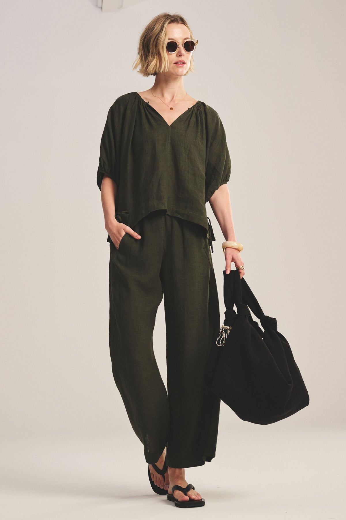 A woman wears the Velvet by Graham & Spencer COSETTE LINEN TOP in olive green with a deep V neckline, paired with matching bottoms, sunglasses, sandals, and a large black bag, set against a plain background.-40808162361537