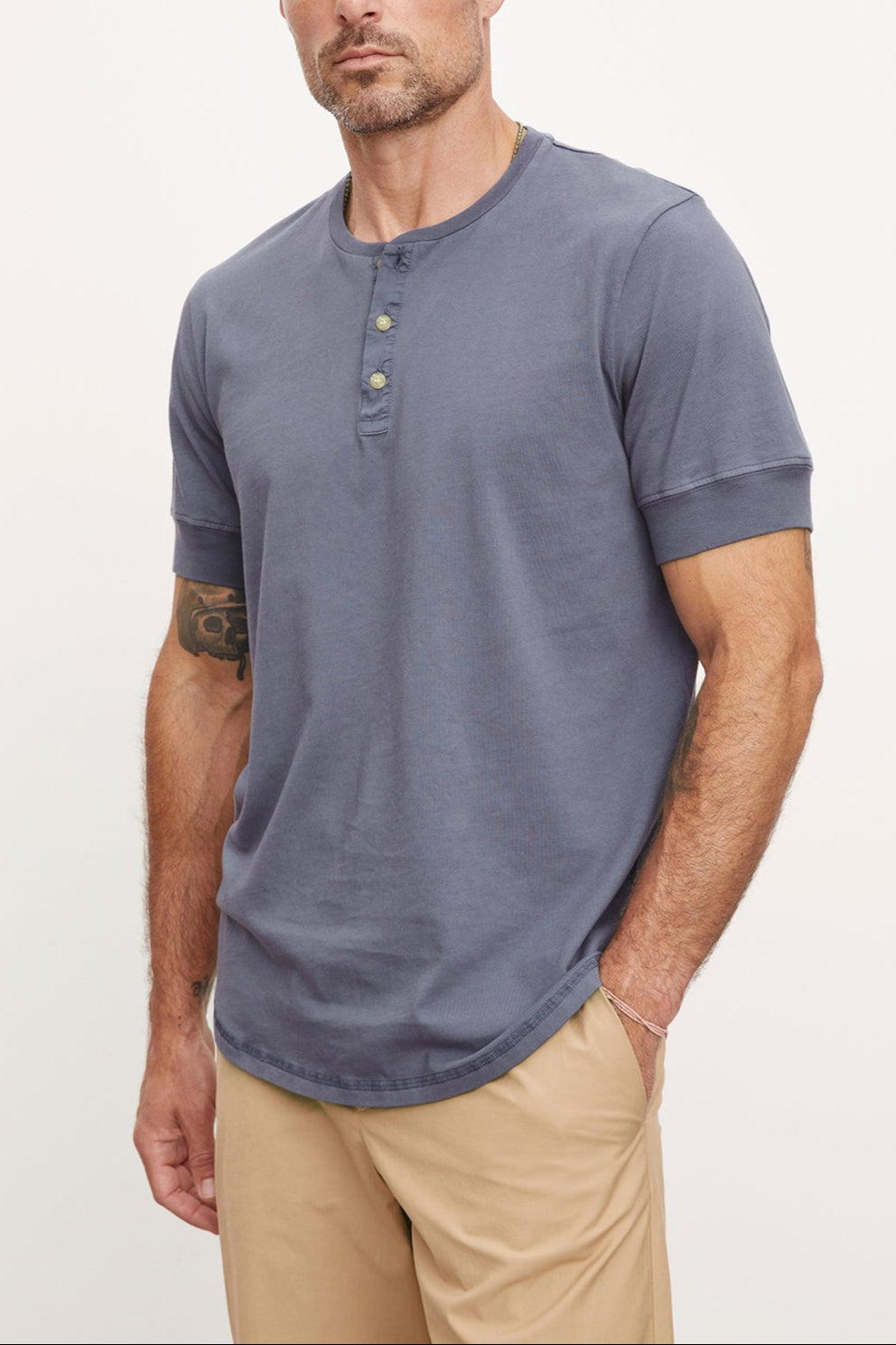 A man with a beard and tattoo on his arm, wearing a gray DEON HENLEY shirt with Henley details and beige pants, stands against a white background, looking at the camera.-36918611935425
